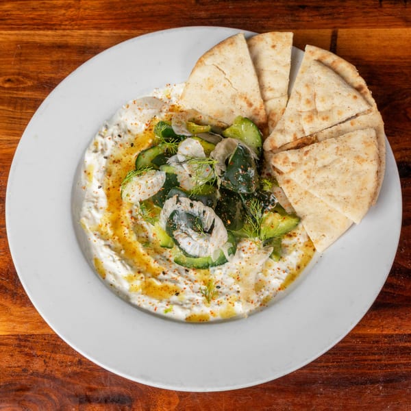 White plate with tzatziki, cucumber salad, and pita bread on a wooden table.