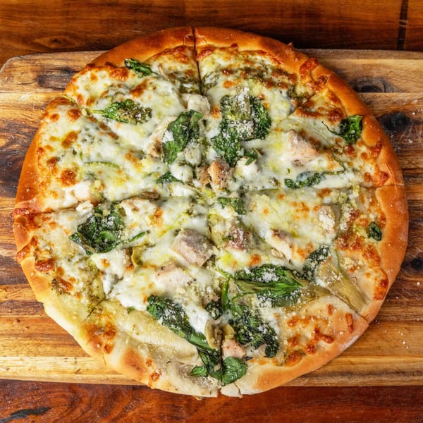 A whole pesto chicken pizza with spinach, artichoke, and cheese on a wooden board.