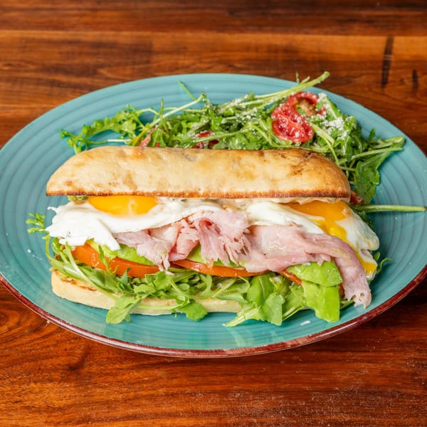 Ciabatta sandwich with ham, fried eggs, avocado, tomato, and arugula, served with a side salad.