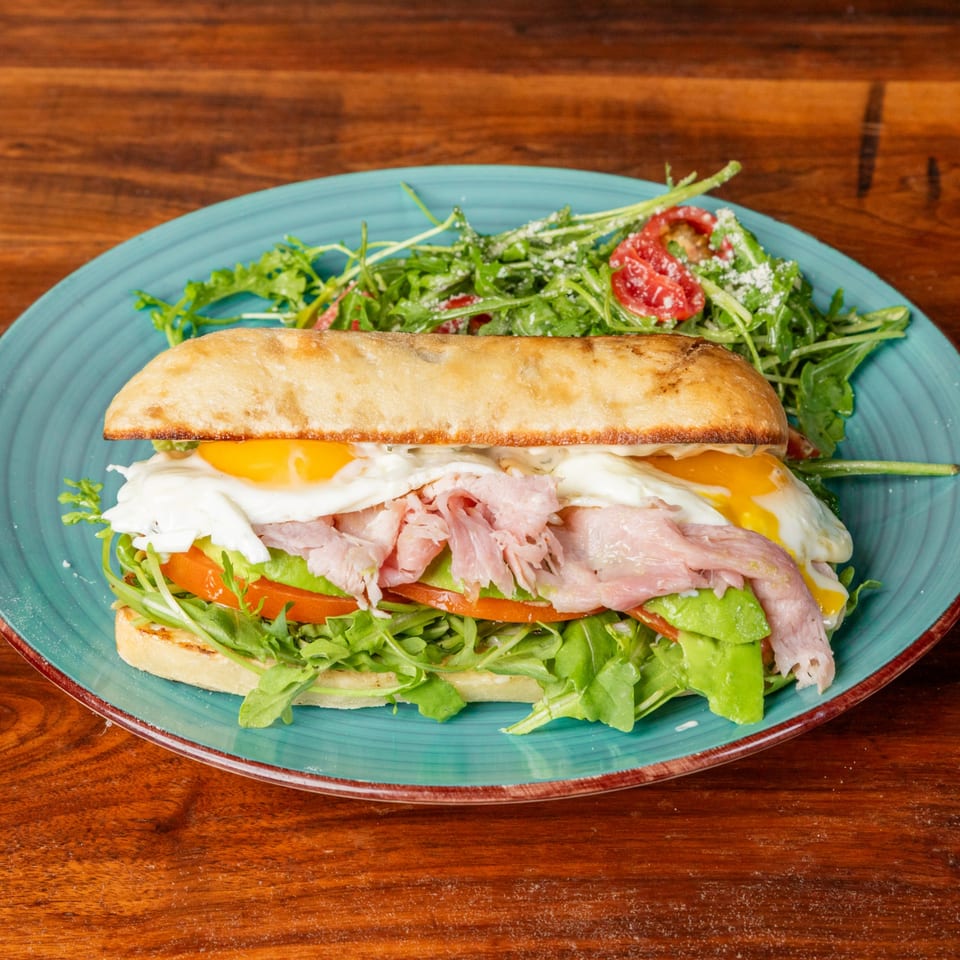 Ciabatta sandwich with ham, fried eggs, avocado, tomato, and arugula, served with a side salad.